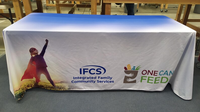 May include: A white tablecloth with a blue gradient at the top. The tablecloth features a superhero child, the text "IFCS Integrated Family Community Services", and the text "ONE CAN FEED" with a graphic of a hand holding a grocery bag.