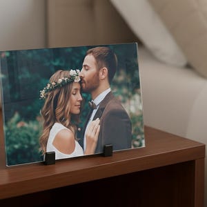 Op de afbeelding: Een rechthoekige fotoprint op glas met een koppel in een bosrijke omgeving, waarbij de persoon links een bloemenkrans en wit topje draagt en de persoon rechts een bruin pak. De print wordt vastgehouden door twee zwarte clips.