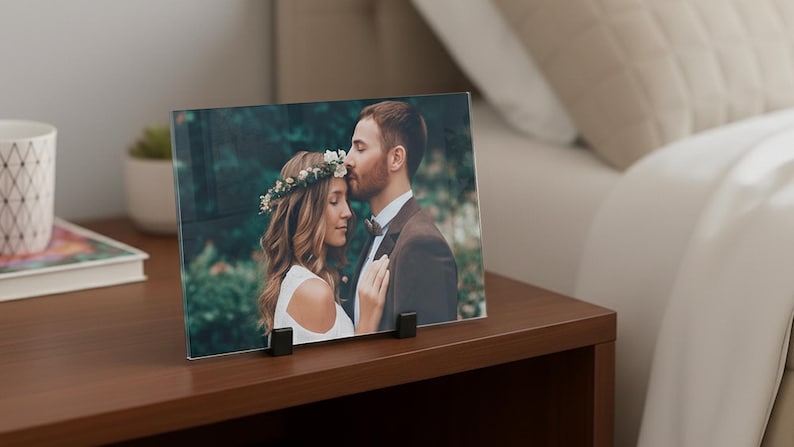 Op de afbeelding: Een rechthoekige fotoprint op glas met een koppel in een bosrijke omgeving, waarbij de persoon links een bloemenkrans en wit topje draagt en de persoon rechts een bruin pak. De print wordt vastgehouden door twee zwarte clips.