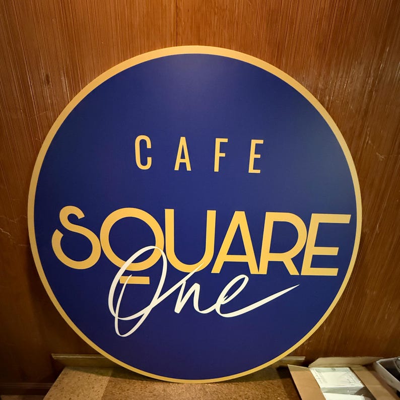 May include: A round, blue and gold sign with the text "Cafe Square One" in a stylized font.