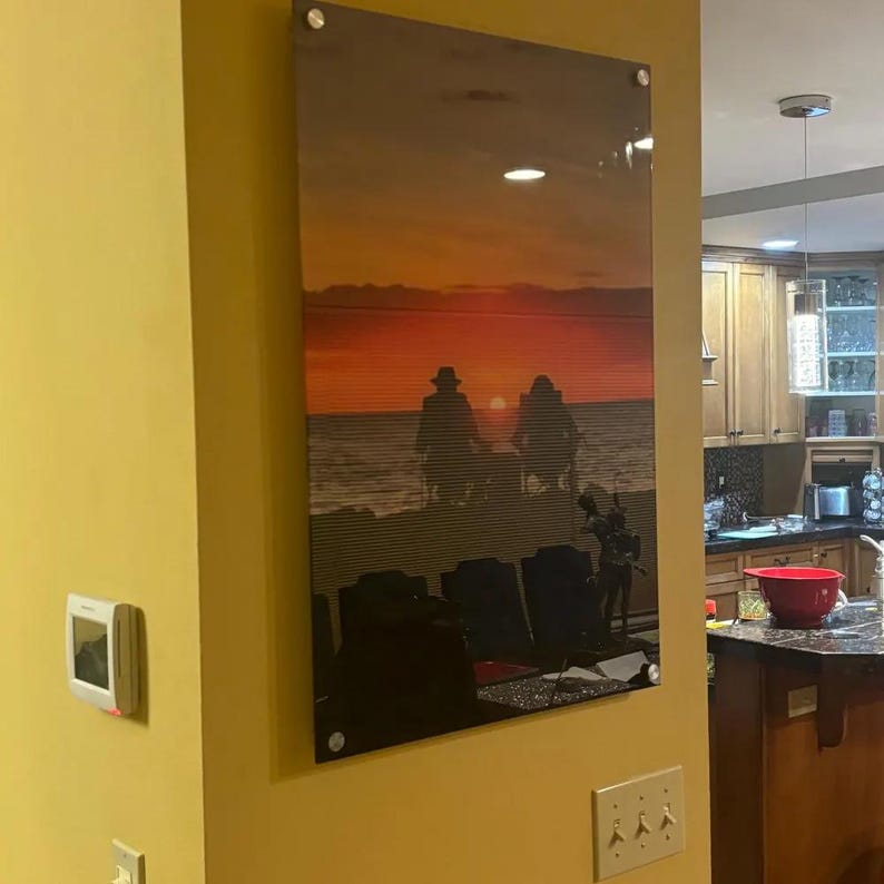Op de afbeelding: Een rechthoekige acrylprint aan de muur met een zonsondergang boven de oceaan en twee silhouetten van figuren die op stoelen zitten. Het kunstwerk is aan de muur bevestigd met vier zilverkleurige metalen afstandhouders.