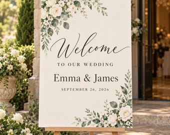 Op de afbeelding: Een rechthoekig welkomstbord voor een bruiloft op een houten ezel met witte rozen en groen blad in de hoeken. De tekst luidt Welcome TO OUR WEDDING Emma & James SEPTEMBER 26, 2026 in zwarte letters.