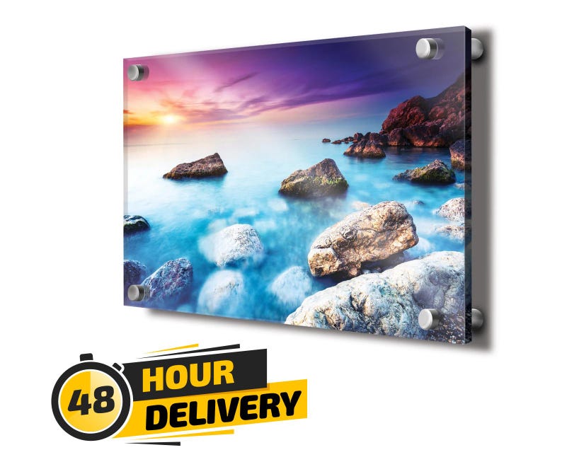 Op de afbeelding: Een rechthoekige acryl wandprint met een zonsondergang boven een rotsachtige kustlijn met blauw water en paarse wolken. De print is bevestigd met vier zilveren metalen afstandhouders op de hoeken. Grafische tekst 48 HOUR DELIVERY.
