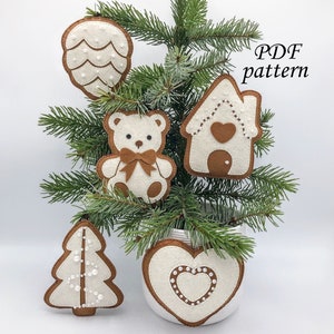 May include: Five felt Christmas decorations in the shape of a pine cone, a house, a teddy bear, a Christmas tree, and a heart. The decorations are white with brown outlines and decorated with white beads. The decorations are hanging on a small artificial Christmas tree.