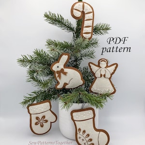 May include: Five felt Christmas ornaments in the shape of a candy cane, bunny, angel, mitten, and stocking. The ornaments are white with brown outlines and are decorated with white beads. The ornaments are displayed on a small artificial Christmas tree.