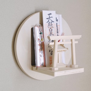 May include: A small wooden torii gate shrine with a white roof and a shelf for displaying decorative items. The shelf is attached to a round wooden plaque with the Japanese characters "天照皇大神" written on it.