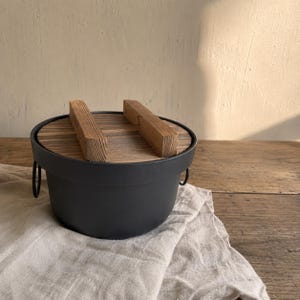 May include: A round, matte black cooking pot with a flat, circular wooden lid. The lid features a visible wood grain pattern and two parallel, rectangular wooden handles on top. The pot has small, circular metal side handles on each side.