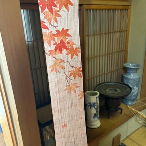 Peut inclure: Une longue et étroite bande de tissu crème suspendue avec un motif de feuilles d'érable rouges. Le tissu a une surface texturée et est frangé en bas. Le tissu est suspendu dans une pièce aux murs et à la porte en bois.