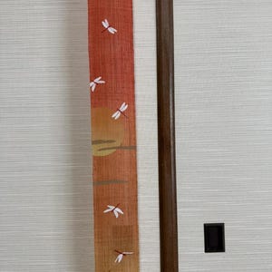 以下が含まれることがあります： 夕焼けと白いトンボが描かれた、細長い布のタペストリー。オレンジ、イエロー、ブラウンのグラデーションで、下部に細い黒い線が引かれている。木製のフレームで壁に飾られている。