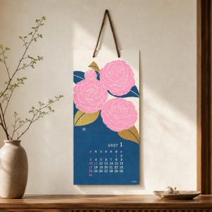 May include: A vertical wall calendar for January 2027 featuring a dark blue background with three large pink camellia flowers and gold-coloured leaves. The calendar is suspended by a dark brown leather strap and displays the month and dates.