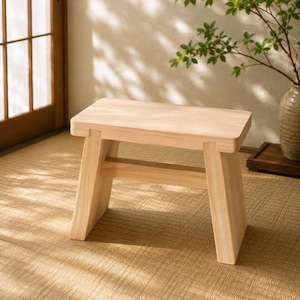 May include: A small, rectangular wooden stool with a light natural finish. The stool features four sturdy, slightly angled legs connected by a horizontal support beam, providing a stable base and a smooth, flat top surface for seating or display.