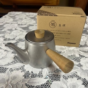 May include: A silver teapot with a wooden handle and lid. The teapot is sitting on a white lace tablecloth. The box behind the teapot is brown with Japanese characters and the word "茶" (tea) in a circle.