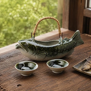 May include: A ceramic teapot shaped like a fish with a dark green and cream gradient finish, featuring a woven rattan handle. Two matching small ceramic bowls with a dark green interior and cream exterior are positioned in front of the teapot.