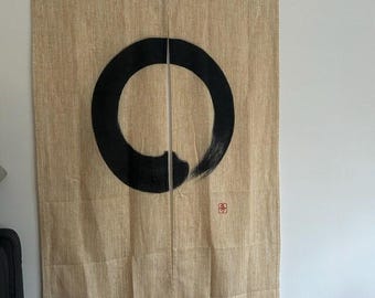 May include: A rectangular beige fabric curtain featuring a large, hand-painted black ink circle in the centre. The fabric has a visible woven texture. A small red square stamp with black characters is positioned below the right side of the circle.