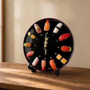 May include: A round black wall clock featuring twelve miniature sushi pieces as hour markers. The clock face displays gold Japanese kanji numerals and ornate gold hands. It is supported by a black plastic stand, showcasing a glossy finish.