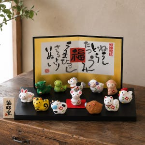 May include: A set of twelve small ceramic animal figurines representing the Chinese zodiac, arranged on a black rectangular base in front of a gold-colored folding screen with black Japanese calligraphy and a red square character in the center.