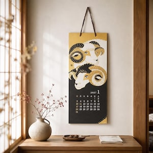 Può includere: Un calendario da parete verticale per gennaio 2027 con un'illustrazione grafica di due arieti con corna ricurve in nero, bianco e oro. Il calendario è appeso a un cordino scuro e mostra il mese, l'anno e una griglia con le date.