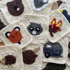 May include: A collection of cream-colored crochet squares, each featuring a different woodland animal or nature-themed design. Animals include a bear, fox, raccoon, and moose. Other squares depict a campfire, leaf, and a camper. The squares are arranged on a wooden surface.