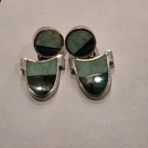 May include: A pair of silver-toned earrings with a geometric design. Each earring features a round top and a shield-shaped bottom, both inlaid with green and black stone. The earrings are displayed on a light-colored surface.