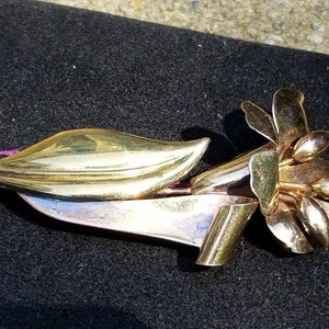 May include: A gold-toned floral brooch with a stylized flower and leaf design. The brooch has a polished finish and is set against a dark background. The brooch is a vintage piece.