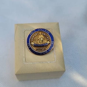 May include: A gold-toned Pace College Nursing pin with blue and white enamel, set on a beige box. The pin features text around the edge and a central image of a figure. The box has a silver border.