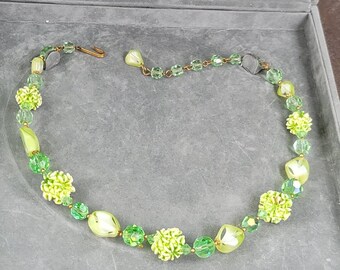 May include: A vintage necklace featuring a variety of green beads and accents. The necklace includes round, faceted, and floral-shaped beads in shades of light and dark green. A gold-toned clasp secures the necklace.