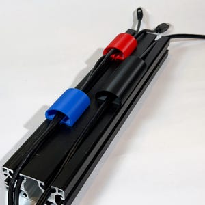 Könnte beinhalten: Ein schwarzer Kabelorganisator aus Metall mit drei farbigen Kabelclips: blau, rot und schwarz. Die Clips halten mehrere schwarze Kabel ordentlich angeordnet. Der Organizer liegt auf einer weißen Oberfläche und sorgt für ein sauberes und organisiertes Aussehen.