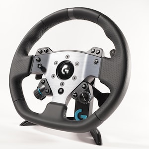 May include: A black and silver racing wheel with a perforated leather grip. The wheel has multiple buttons, dials, and a central logo. The wheel is mounted on a black stand, and the overall design is sleek and modern.