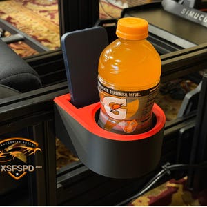 Puede incluir: Un portavasos y un soporte para teléfono negros y rojos sujetos a un marco de metal negro. Una botella de bebida deportiva naranja y un teléfono inteligente negro están en los soportes. El logotipo "RACERBIKE SPEED" es visible.