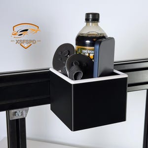 May include: A black storage container with a white rim, holding a bottle of beverage, a smartphone, and two black discs. The container is mounted on a black metal frame. The logo "XFSFPD" is visible in the upper left corner.