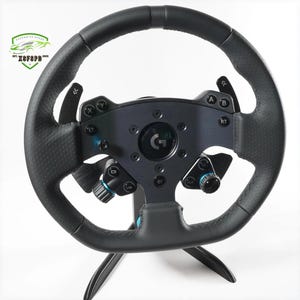 以下が含まれることがあります： 黒とグレーのレーシングホイール。テクスチャード加工されたグリップと複数のボタンとダイヤルを備えています。 ホイールの中央には「G」の文字があり、黒いスタンドに取り付けられています。 このホイールはビデオゲームで使用するように設計されています。