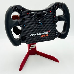 Puede incluir: Un volante de carreras McLaren GT3 negro con botones rojos, verdes y azules, un diseño de fibra de carbono negro y blanco y un soporte rojo con detalles blancos.