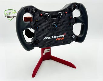 Könnte beinhalten: Ein schwarzes McLaren GT3-Rennlenkrad mit mehreren Knöpfen und Bedienelementen, montiert auf einem roten Ständer. Das Lenkrad hat eine Kohlefaserstruktur und das McLaren-Logo. Der Ständer hat ein dreibeiniges Design.