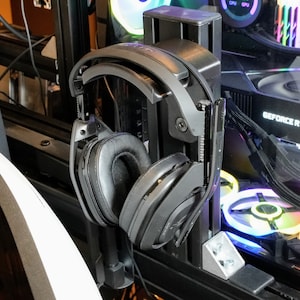 May include: A black headset with a padded headband and earcups is hanging on a black metal stand. The headset is attached to the stand with a metal clip. The stand is mounted on a computer case with a black and white design.