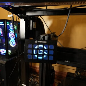 May include: A black Stream Deck with a blue backlit button showing a play button icon. The Stream Deck is mounted on a black metal stand.