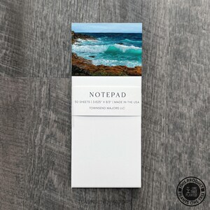 May include: A white notepad with a scenic ocean image at the top. The notepad has the word "NOTEPAD" printed on it, along with the text "50 SHEETS | 3.625" x 8.5" | MADE IN THE USA". The notepad is on a wooden surface.