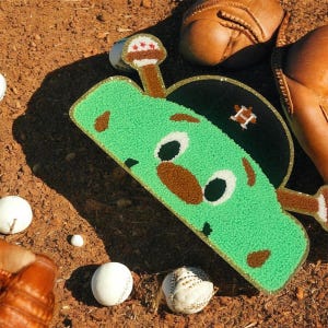 Może przedstawiać: Zielony, kreskówkowy przedmiot w kształcie kija do baseballu z twarzą, złotym obramowaniem i czarną czapką baseballową z logo "H". Piłki do baseballu i rękawica do baseballu otaczają przedmiot.