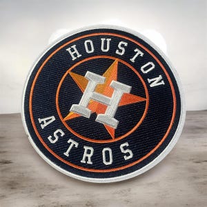 Może przedstawiać: Okrągła haftowana naszywka z logo Houston Astros. Wzór zawiera granatowe tło z pomarańczową gwiazdą, srebrną literą H oraz białym napisem HOUSTON ASTROS ułożonym w okrągły wzór wokół środka naszywki.