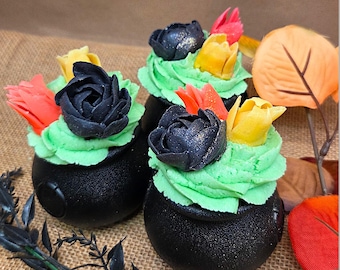 May include: Three black cauldron-shaped treats topped with green frosting and colorful flower decorations. The flowers are in shades of black, yellow, and red. The treats are arranged on a burlap surface, with fall-themed decorations.