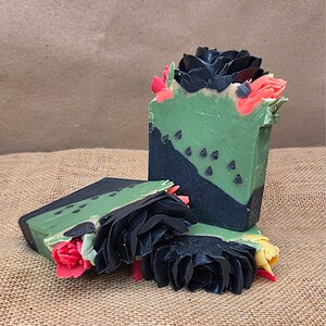 May include: Handmade soap bars with a layered design. The soap features a green top layer with black accents and decorative black floral and red accents. The soap bars are arranged on a textured, neutral-toned surface.