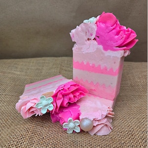 May include: Handmade pink and white soap bars with decorative floral accents. The soap bars feature layers of pink and white, topped with sculpted pink flowers and small white flower embellishments. The soap bars are displayed on a textured brown surface.