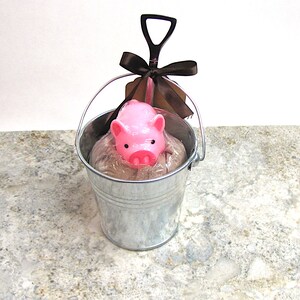 May include: A small pink pig figurine in a metal bucket with a brown ribbon and a small shovel. The pig is sitting in a bed of brown material.