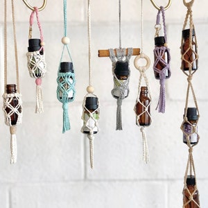 Może przedstawiać: Dziewięć makramowych uchwytów na butelki z frędzlami wiszących na białej ceglanej ścianie. Każdy uchwyt wykonany jest z naturalnego sznurka i zawiera małą szklaną butelkę. Butelki są wypełnione olejkami eterycznymi.