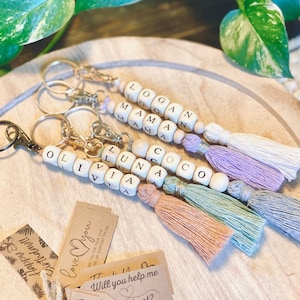 May include: Four wooden bead keychains with tassels. Each keychain has a different name spelled out on the beads: Logan, Mama, Olivia, and Luna. The tassels are in shades of white, lavender, peach, and light green.