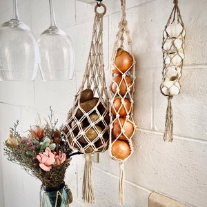 Pode incluir: Três organizadores pendurados em macramé com diferentes conteúdos. O primeiro organizador contém cebolas, o segundo batatas e o terceiro bulbos de alho.