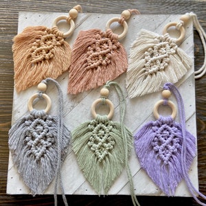 May include: Six handmade macrame leaf ornaments with wooden rings. The leaves are in various colors, including beige, pink, white, gray, green, and purple.