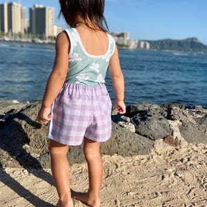 May include: A child in a light green vest top with white floral patterns and purple plaid shorts. The shorts have a white stripe down the side. The child is on a sandy beach with the sea in the background.