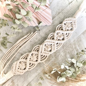 May include: A beige macrame belt with a diamond pattern. The belt is made of cotton cord and is tied with a knot at the end.