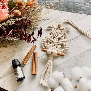 May include: A white macrame owl wall hanging with wooden beads. The owl is hanging from a white cord. There are cinnamon sticks and a bottle of essential oil on a white wooden surface.