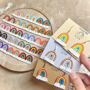 May include: A variety of rainbow-colored crocheted earrings with white fringe. The earrings are displayed on a wooden plate and on cards with the text "Island RM Handmade with Aloha".